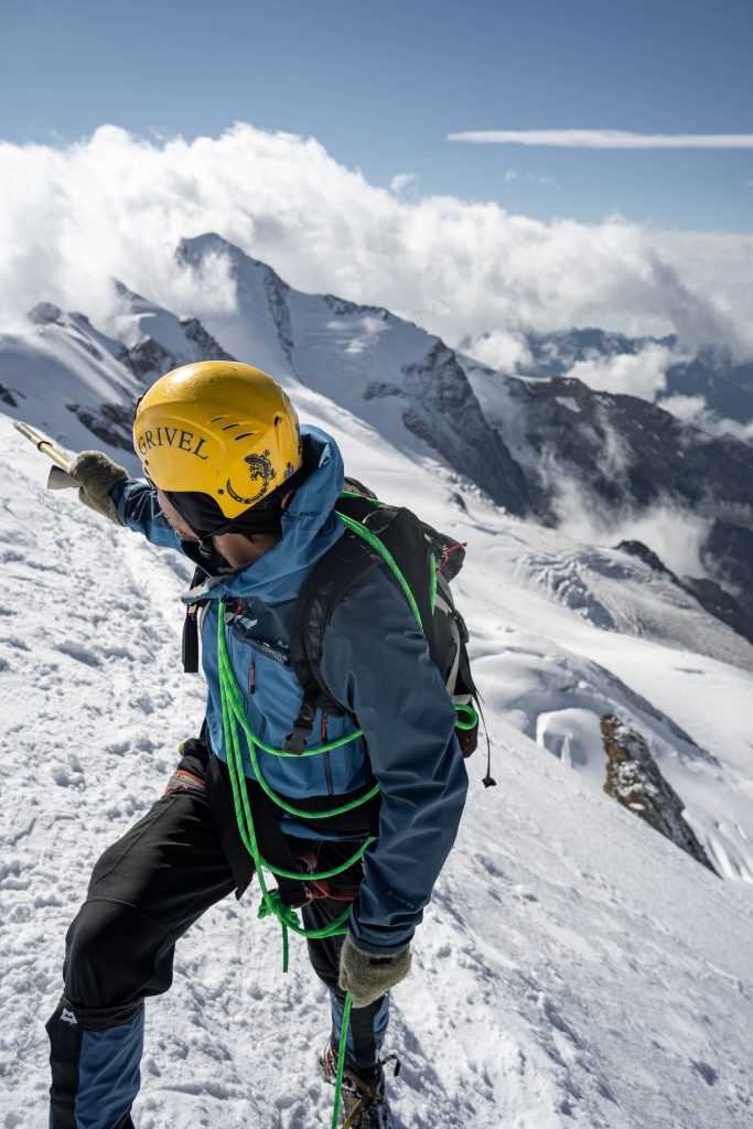 Schönstes Wetter, imposante Landschaft und beste Laune. Mann in blauer Jacke am Gipfelgrat eines vergletscherten Berges.